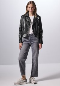 Giubbotto da biker in pelle nera con chiusure a zip, abbinato a jeans ampi grigi chiari e sneakers beige. Maglietta bianca semplice sotto.