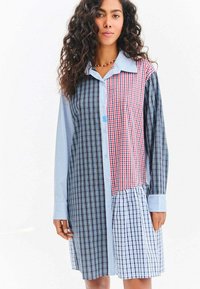 Žena s kudrnatými vlasy nosící dlouhý rukáv s patchworkovým kostkovaným šatem s modrými, červenými a černými vzory, stojící před bílým pozadím.