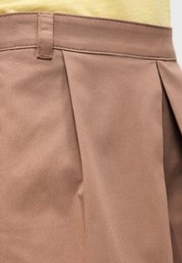 Κοντινή λήψη καφέ παντελονιού με πιέτες και θηλιά για ζώνη, σε συνδυασμό με κίτρινο τοπ.