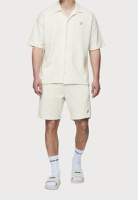 Uomo che indossa una camicia a maniche corte di colore bianco sporco con texture e pantaloni corti abbinati, con calze bianche e sandali scivolosi su sfondo bianco.