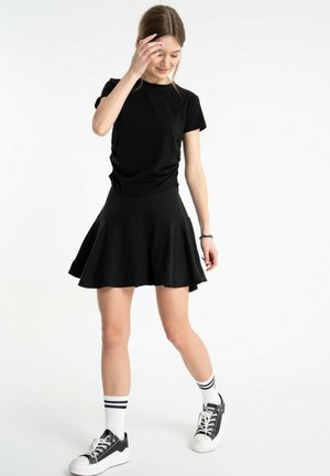 Młoda kobieta ubrana w czarną koszulkę, czarną rozkloszowaną spódnicę, białe skarpetki z czarnymi paskami i czarne trampki, stojąca na białym tle.