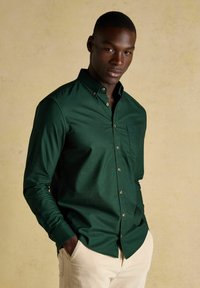 Bărbat purtând o cămașă verde închis cu nasturi și buzunar la piept și pantaloni bej, stând în fața unui fundal bej simplu.