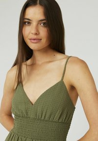 Robe vert olive en tissu texturé et froissé. Dotée d'un décolleté en V, de fines bretelles et d'une taille élastique froncée.