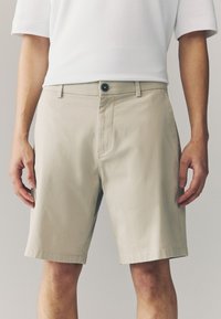 Beežid lühikesed püksid, millel on sile tekstuur, esiküljel nööpidega sulgemine, vöökohad ja küljetaskud. Püksid on sirge lõikega ja põlvepikkused.