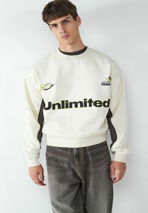 Crème sweatshirt met "Unlimited" in vetgedrukte zwarte tekst, voorzien van kleine logo's in groen en blauw. Donkere accenten op de zijpanelen en geribde manchetten.