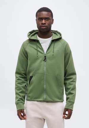 Homme portant un sweat à capuche vert zippé avec cordons et pantalon de couleur claire, debout devant un fond clair uni.
