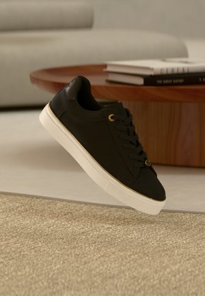 Czarny niski sneaker z białą podeszwą i złotymi oczkami unoszący się nad beżowym dywanem obok drewnianego stolika kawowego z książkami.