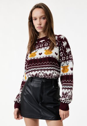 Vrouw draagt een bordeauxrode en witte kersttrui met gele rendiergezichten en een zwarte leren minirok, staand tegen een witte achtergrond.