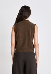 Brauner ärmelloser Strickpullover mit gerippter Schulter- und Saumdetails. Getragen mit dunklen, taillierten Hosen. Weiche Textur und figurbetonte Form.