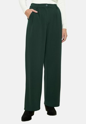 Pantalons larges en tissu vert foncé avec plis à l'avant, passants de ceinture, poches latérales et fermeture à boutons, associés à des chaussures noires.