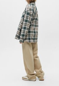 Persoon die zijwaarts staat in een oversized groen en beige geruite shirt, losse beige broek en witte sneakers tegen een effen achtergrond.