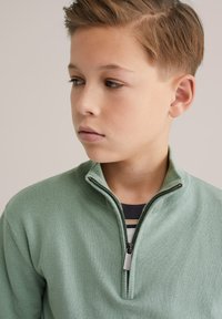 Pull-over vert menthe avec un col montant et une fermeture éclair. Tissu en maille douce, avec des accents côtelés. Texture unie sans motifs apparents.