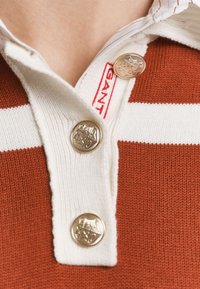 Polo shirt en tricot avec un corps orange rouille et des rayures blanches, doté d'un col, de trois boutons dorés et d'une étiquette rouge.