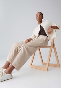 Femme portant un pantalon beige à jambes larges, un haut noir, un cardigan blanc et des baskets blanches, allongée sur une chaise pliante en bois contre un fond uni.
