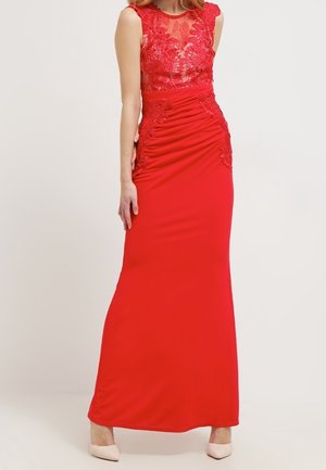 Femme portant une longue robe rouge avec corsage en dentelle et détail froncé à la taille, associée à des chaussures à talons hauts beige clair.