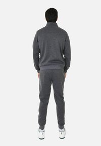 Tuta in felpa grigia con colletto alto, polsini e cintura a costine. I pantaloni sono slim fit. Le scarpe presentano accenti bianchi e verdi.
