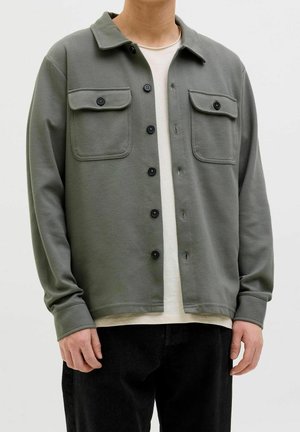 Personne portant une veste-chemise grise à boutons avec deux poches poitrine et des boutons noirs sur un t-shirt blanc et un pantalon noir.