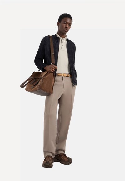 Chaqueta de ante oscura con cremallera sobre una camiseta polo clara, pantalones beiges, cinturón de cuero marrón y bolso de ante marrón con zapatillas marrones.