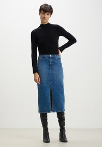 Pullover a collo alto nero a costine, abbinato a una gonna di denim blu di media lunghezza con uno spacco frontale. Stivali neri alti fino al ginocchio con tacco completano il look.