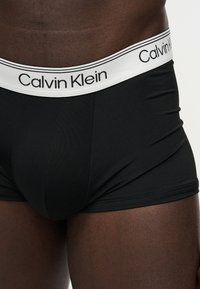 Ανδρικός κορμός που φοράει μαύρα μποξεράκια Calvin Klein με λευκή μέση με μαύρο λεκτικό λογότυπο.