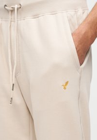 Μπεζ παντελόνι φόρμας με ριμπ ζώνη μέσης, κορδόνι και πλαϊνές τσέπες. Διαθέτει μικρό κίτρινο κεντητό λογότυπο πουλιού στο αριστερό πόδι.