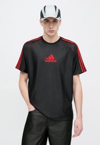 Czarny t-shirt z krótkim rękawem z czerwonym logo Adidas i paskami, zestawiony z teksturowaną szarą czapką z białym logo. Prosty design i sportowy styl.