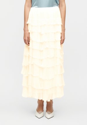 Γυναίκα που φοράει κρεμ μάξι φούστα με στρώσεις βολάν και μυτερά λευκά τακούνια, στέκεται μπροστά σε απλό ανοιχτό φόντο.