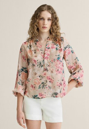 Kvinde med krøllet hår iført en beige blomsterbluse med opklappede ærmer og hvide shorts, stående mod en ensfarvet baggrund.