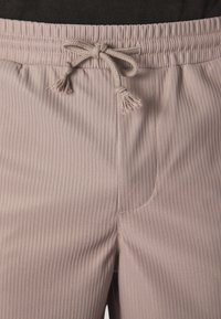 Pantalons de jogging côtelés rose pâle avec une taille élastique, cordon de serrage réglable et poche latérale. Texture douce avec des détails de couture visibles.