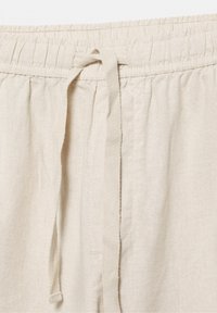 Prim-plan al taliei pantalonilor bej din in, cu elastic și șnur din material legat în față, în centru.