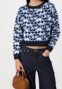 Sweter dzianinowy z niebieskim wzorem serca na granatowym tle, krótki fason, z ściągaczami; w zestawieniu z ciemnymi dżinsami. Brązowa torebka.