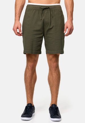 Shorts verdes oliva con cintura elástica y cordón. Tejido suave, hasta la rodilla, con dobladillo doblado, combinados con zapatillas oscuras.