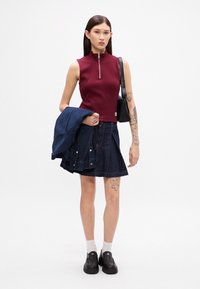 Haut sans manches bordeaux avec une fermeture éclair, associé à une jupe en denim bleu foncé. Veste sombre portée sur le bras, chaussures noires à enfiler, chaussettes blanches.
