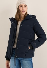 Marinblått puffjacka med dragkedja framtill, knapplåsningar och huva. Har horisontella quiltade linjer och en logotyp på ärmen.
