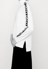 Vit polotröja med svart DKNY-logotyp längs ärmarna. Har sidokilar och en mjuk textur.