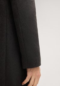 Gros plan sur la main d'une personne reposant à côté de la manche d'un manteau en laine texturée sombre sur un fond clair.