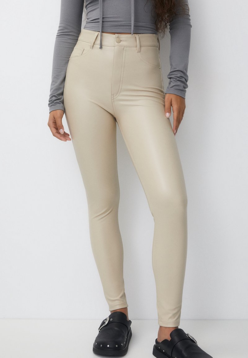 Leggings beiges taille haute en matériau extensible et lisse. Comprend un bouton à l'avant et des poches. Portés avec des sabots noirs.