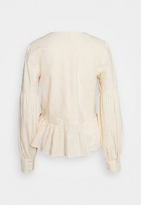 Blusa color crema con strisce testurizzate, maniche a sbuffo e bordo a peplum. Presenta una scollatura a V nella parte posteriore e polsini con bottoni.