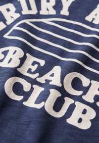 Granatowy materiał z białymi teksturowanymi literami napisanymi "BEACH CLUB" oraz grafiką deski surfingowej, charakteryzującą się gładkimi liniami i swobodnym stylem.