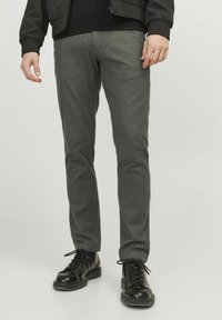Muž v šedých slim-fit kalhotách a černých šněrovacích botách, stojící na jednolitém světlém pozadí.