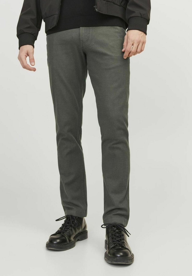 Muž v šedých slim-fit kalhotách a černých šněrovacích botách, stojící na jednolitém světlém pozadí.