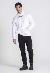 Sudadera blanca con capucha y logo negro, combinada con jeans negros y zapatillas negras, con un ajuste slim y una textura de tela suave.