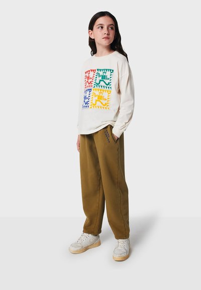 Beige Langarmshirt mit buntem geometrischem Tierdruck. Olivgrüne Jogginghose mit blauen Akzenten. Weiße Sneakers mit grauen Details.