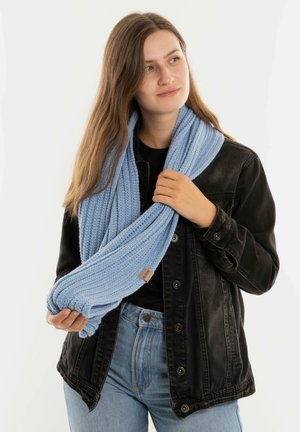 Jonge vrouw in een zwarte denimjas en een lichtblauwe gebreide sjaal, die de uiteinden van de sjaal vasthoudt en iets naar rechts kijkt, op een witte achtergrond.
