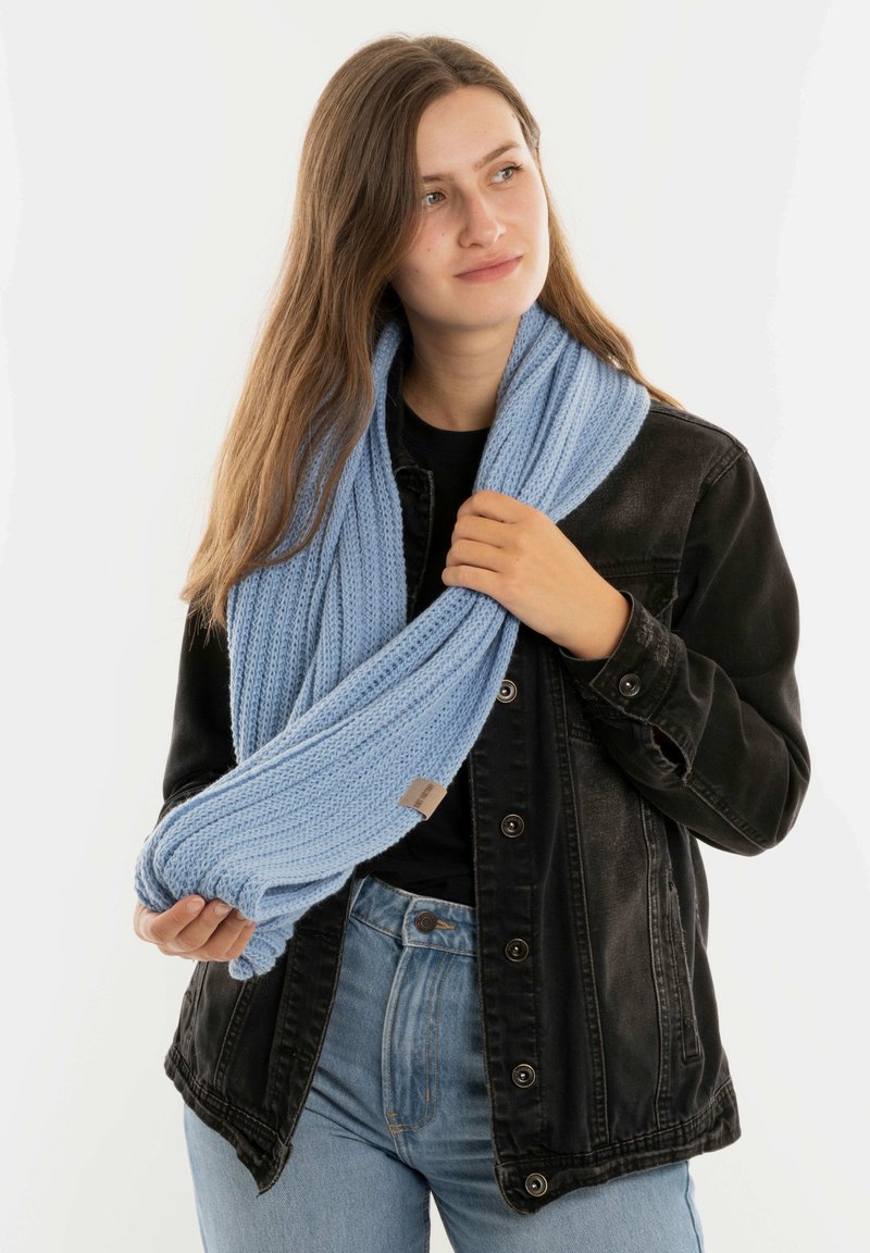 Jonge vrouw in een zwarte denimjas en een lichtblauwe gebreide sjaal, die de uiteinden van de sjaal vasthoudt en iets naar rechts kijkt, op een witte achtergrond.