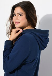 Felpa blu navy con cappuccio e polsini a coste; tessuto morbido; mano del modello vicino al colletto. Design minimalista su sfondo bianco.