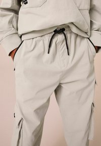 Pantalons cargo gris clair avec taille élastique et cordon de serrage noir ; comporte des poches latérales et des coutures ton sur ton. Texture lisse et coupe décontractée.