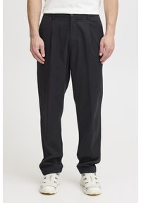 Black, pleated trousers made from lightweight fabric with a relaxed fit, paired with white and navy sneakers. Flat front and button closure.