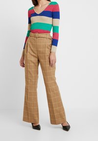 Pasiasty sweter dzianinowy w kolorach czerwonym, niebieskim, zielonym i beżowym; zestawiony z wysokim stanem beżowych spodni w kratę z żółtym wzorem w kratkę i czarnymi szpilkami.