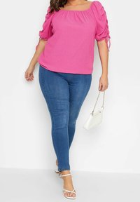 Růžová texturovaná blůza s nabíranými rukávy a šňůrkami, doplněná modrými přiléhavými džíny. Bílá kabelka a sandály na pásky dotvářejí vzhled.